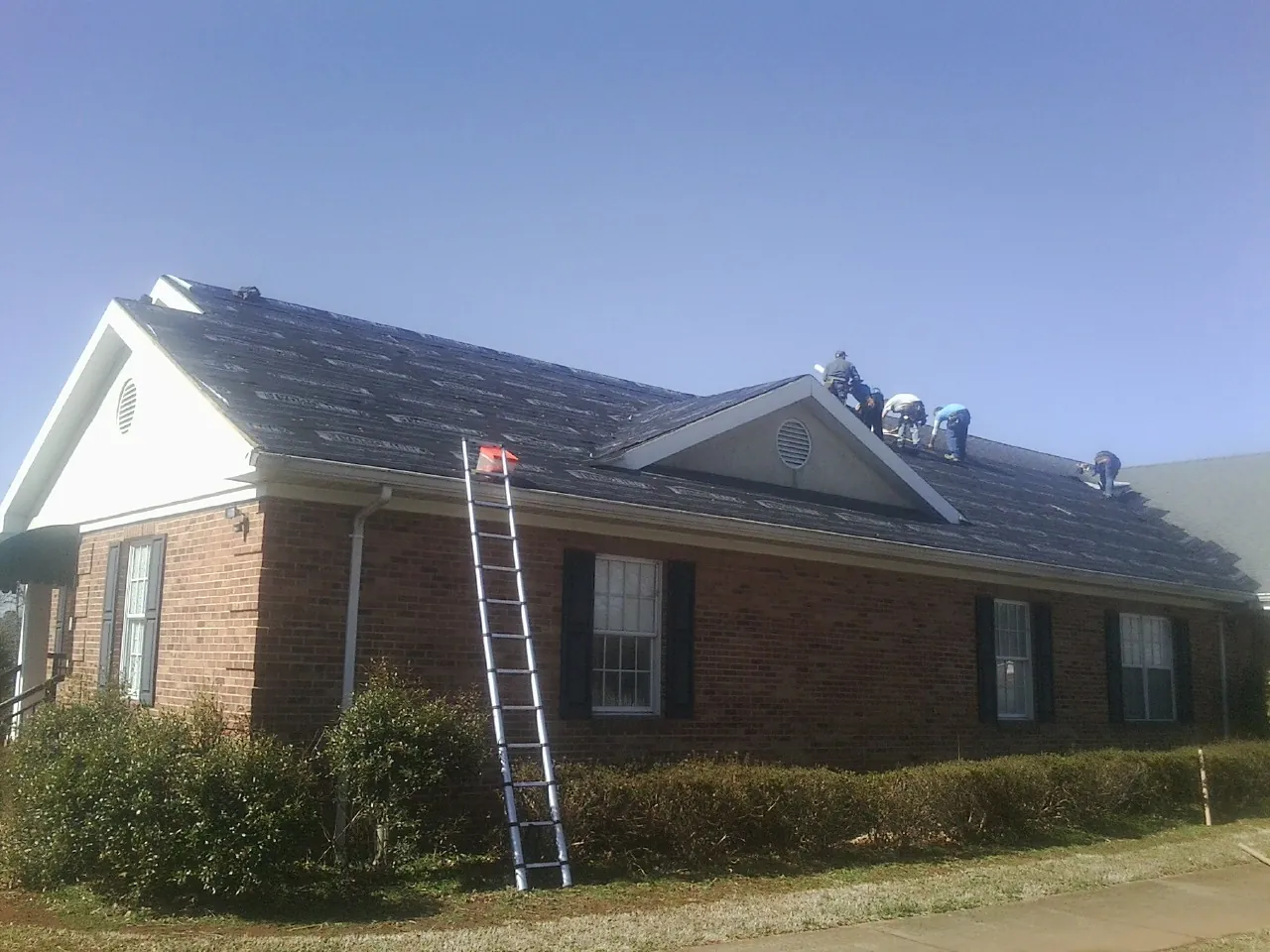 Crew tearing shingles off a brick ranch roof with a ladder set — Emergency Roof Repair work by our crew in Wetmore