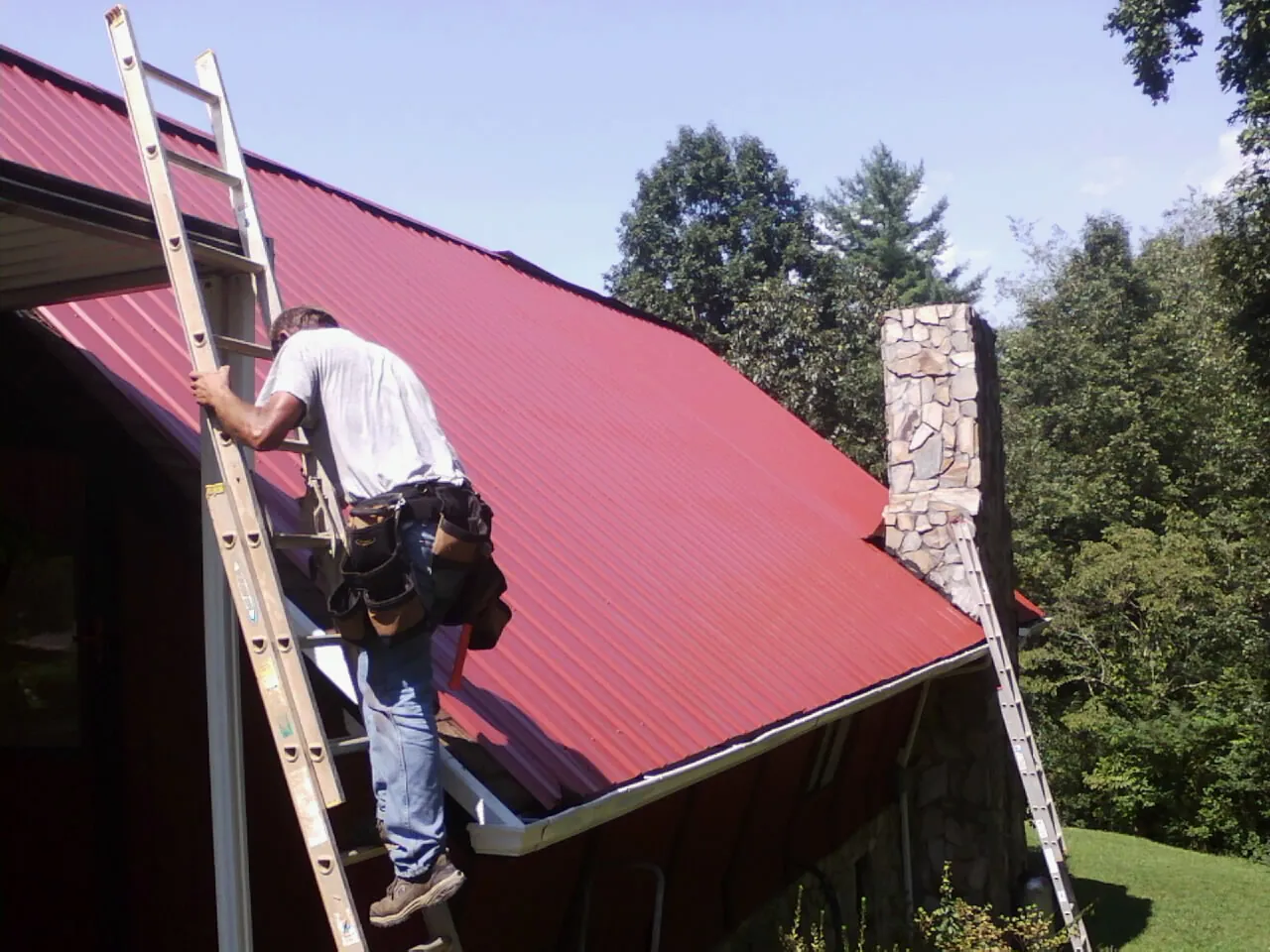 Roofer on a ladder beside a red metal roof and stone chimney — Soffit & Fascia Repair by Roofing Brothers in Wetmore